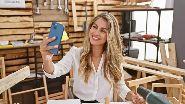 Attractive young blonde woman carpenter confidently snapping a selfie while sitting at carpentry table in her woodworking workshop