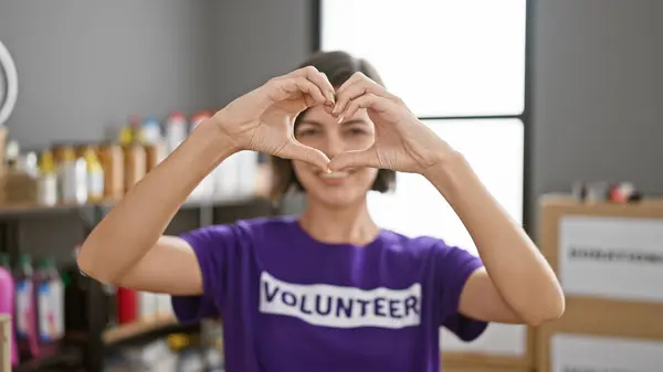 With hands forming a love symbol, a young, beautiful hispanic woman with short hair confidently volunteer at a charity center, her radiant smile reflecting the joy of selfless service.