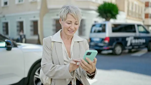 Middle age blonde woman smiling confident using smartphone at street