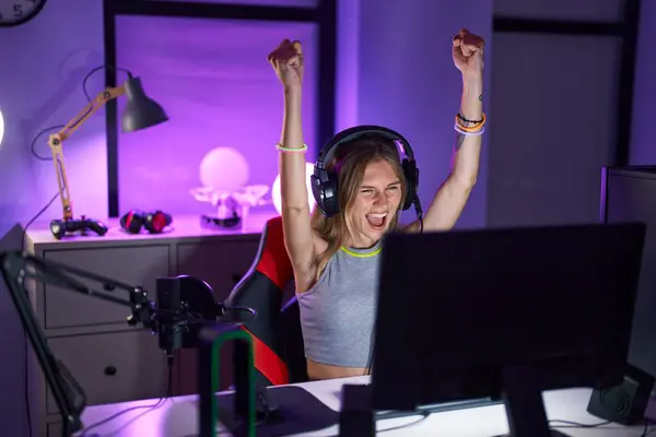 Young blonde woman streamer playing video game with winner expression at gaming room