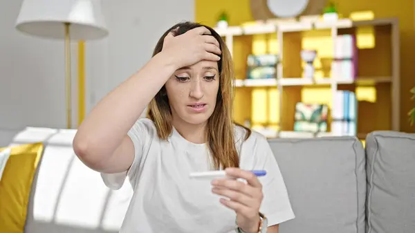 Young blonde woman holding pregnancy test with worried expression at home