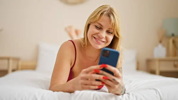 年轻的金发女人穿着性感内衣躺在床上 在卧室里用智能手机 — 图库照片