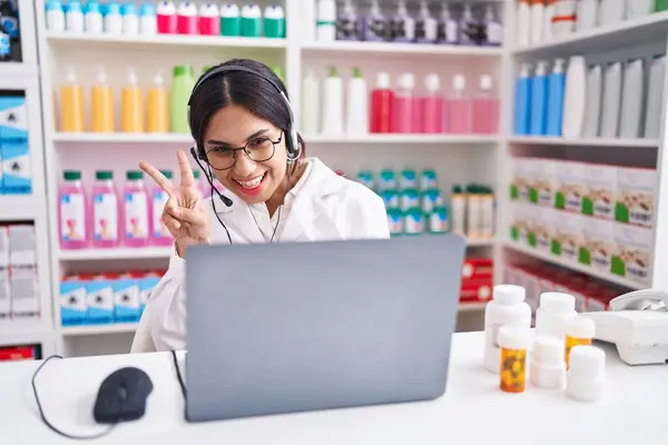 Young arab woman working at pharmacy drugstore using laptop smiling looking to the camera showing fingers doing victory sign. number two.