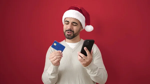 Молодой Латиноамериканец Рождественской Шляпе Помощью Смартфона Кредитной Карты Изолированном Красном — стоковое фото