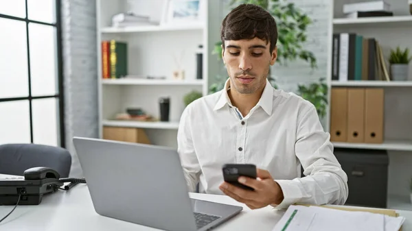 Portrait of a handsome young hispanic business executive, indoors in his office, the serious male worker is busy typing a message on his laptop and texting on his phone
