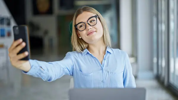 Young blonde woman business worker using laptop making selfie by smartphone at office