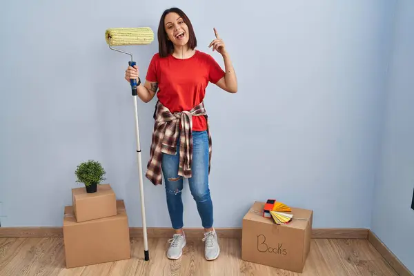 Young hispanic woman painting home walls with paint roller smiling amazed and surprised and pointing up with fingers and raised arms.