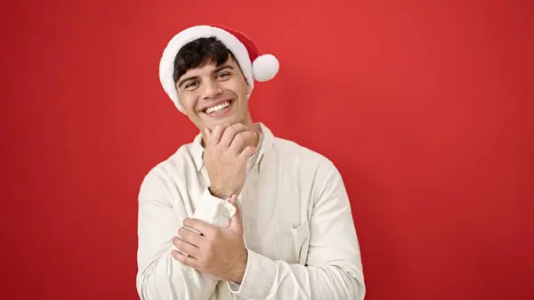 孤立した赤い背景の上にクリスマスの帽子をかぶって自信を持って微笑む若いヒスパニックの男 — ストック写真