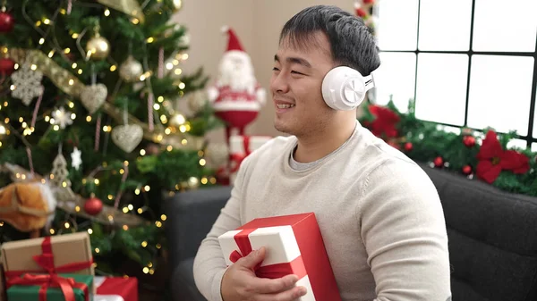 Young chinese man listening to music holding gift by christmas tree at home