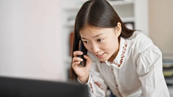 Young chinese woman business worker using computer talking on smartphone at office