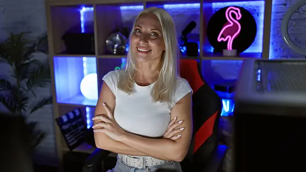 Attractive young blonde streamer plays video game while sitting at home in a dark gaming room, smiling and relaxed. arms crossed, she immerses herself in the virtual world of digital entertainment.