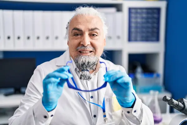 Middle age grey-haired man scientist smiling confident holding security glasses at laboratory