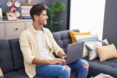 Genç İspanyol adam evdeki koltukta dizüstü bilgisayar kullanıyor.