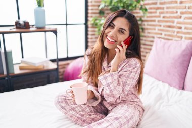 Young beautiful arab woman talking on smartphone drinking coffee at bedroom