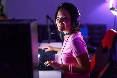 Young hispanic woman streamer playing video game using computer at gaming room