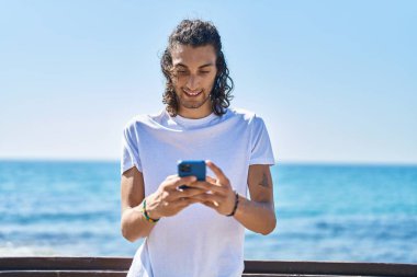 Genç İspanyol adam deniz kenarında akıllı telefon kullanarak gülümsüyor.