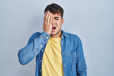 Young hispanic man standing over blue background yawning tired covering half face, eye and mouth with hand. face hurts in pain. 