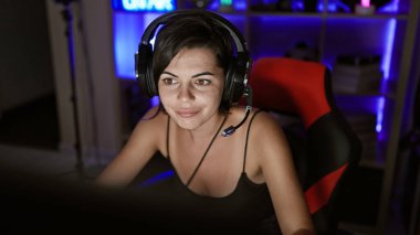 Kendine güvenen genç İspanyol bir kadın, güzel bir flama, oyuna dalmış, karanlık oyun odasında yayılıyor, teknoloji parlıyor, kulaklık takıyor, neşeli bir gülümseme geceyi aydınlatıyor.