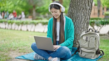 Parkın zemininde dizüstü bilgisayar ve kulaklık kullanan genç Çinli kadın.