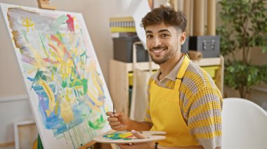 Canlı bir üniversite sanat stüdyosunda ustalıkla gülümseyen kendine güvenen genç Arap sanat öğrencisi, yaratıcılığını fırça ve paletle kullanıyor.