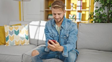 Beyaz bir adam akıllı telefon kullanıyor. Evdeki kanepede oturuyor.