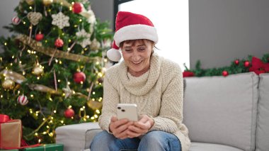 Evde Noel ağacının yanında oturan akıllı telefon kullanan olgun İspanyol kadın.