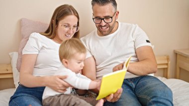 Anne, baba ve bebeğin ailesi yatak odasındaki yatakta oturmuş kitap okuyor.