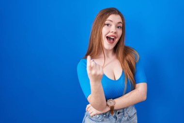 Kızıl saçlı kadın mavi arka planda duruyor. Buraya el kol hareketi yaparak mutlu ve gülümseyen insanları davet ediyor. 