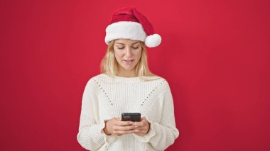 Yılbaşı şapkası takan genç sarışın kadın izole edilmiş kırmızı arka plan üzerinde akıllı telefon kullanıyor.