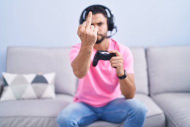 Hispanic young man playing video game holding controller sitting on the sofa showing middle finger, impolite and rude fuck off expression 