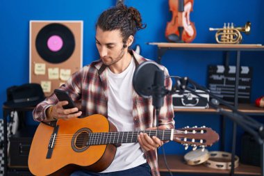 Genç İspanyol müzisyen klasik gitarı müzik stüdyosunda akıllı telefon kullanarak tutuyor.