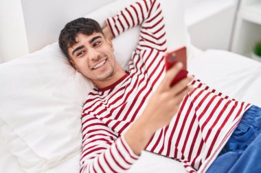 Genç İspanyol adam yatakta akıllı telefon kullanıyor.