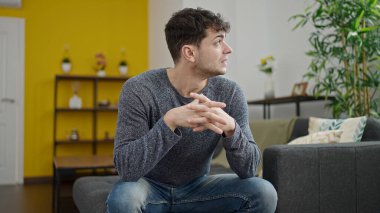 Kanepede oturan Latin asıllı genç bir adam. Şüpheli bir ifadeyle.