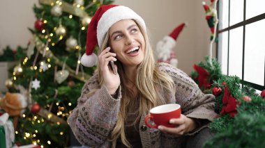 Genç sarışın kadın akıllı telefonda konuşuyor. Evdeki Noel ağacının yanında kanepede kahve içiyor.