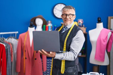 Orta yaşlı, gri saçlı, kendine güvenen bir terzi elbise fabrikasında dizüstü bilgisayar kullanıyor.