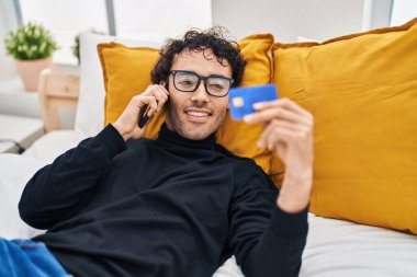 Genç İspanyol adam elinde kredi kartıyla akıllı telefondan konuşuyor. Yatak odasında yatıyor.