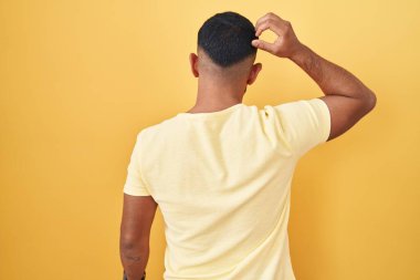 Young hispanic man standing over yellow background backwards thinking about doubt with hand on head 