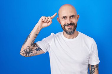 Mavi arka planda dikilen İspanyol dövmeli adam gülümseyerek tek parmağıyla kafasını işaret ediyor, harika bir fikir ya da düşünce, iyi bir hafıza 