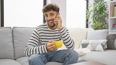 Mutlu Arap adam, rahatlatıcı kahvaltının tadını çıkarıyor, oturuyor, elinde kahve fincanı var. Gülümsüyor, akıllı telefonuyla konuşuyor, rahat evinde..