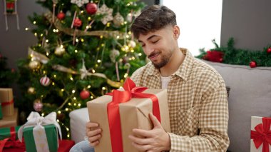 Genç Arap adam elinde hediyeyle evde Noel ağacının yanında oturuyor.