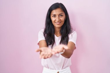 Pembe arka planda duran genç İspanyol kadın avuçlarını birbirine uzatarak gülümsüyor ya da el hareketi yapıyor. Bekleme ve koruma 