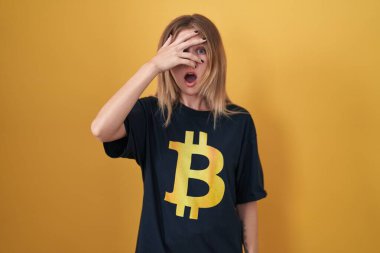 Sarışın, beyaz kadın bitcoin tişört giyiyor. Şoka girmiş, yüzünü ve gözlerini kapamış, utanmış ifadesiyle parmaklarından bakıyor.. 