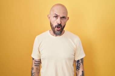 Sarı arka planda duran İspanyol dövmeli adam. Şok suratlı, şüpheci ve alaycı görünüyor. Ağzını açtığında şaşırıyor. 