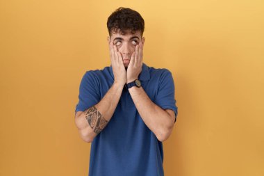 Young hispanic man standing over yellow background tired hands covering face, depression and sadness, upset and irritated for problem 