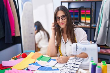 Young beautiful hispanic woman tailor talking on smartphone writing on notebook at clothing factory