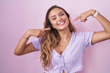 Pembe arka planda duran, neşeli gülümseyen ve parmaklarıyla ve ağzıyla işaret eden genç İspanyol kadın. diş sağlığı kavramı. 