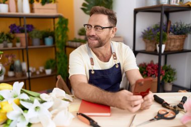 Çiçekçide akıllı telefon kullanarak gülümseyen orta yaşlı bir çiçekçi.