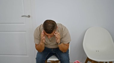 Stresli, yalnız ve hayal kırıklığına uğramış genç bir latin adam, bir sandalyede oturuyor, kendi sorunlarıyla eziyet çekiyor, steril bekleme odasında hüzün havası yayıyor.