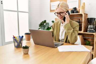 Genç sarışın iş kadını bilgisayarını kullanarak ofiste akıllı telefondan konuşuyor.