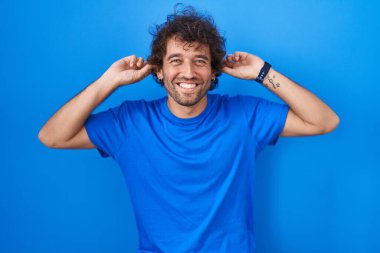 Mavi arka planda dikilen İspanyol genç adam gülümsüyor parmaklarıyla kulaklarını çekiyor, komik bir jest. deneme sorunu 